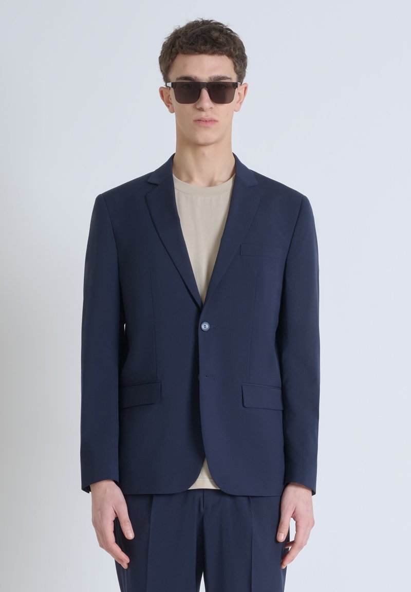 Blazer blu navy con vestibilità slim, due tasche anteriori, revers a scatto e chiusura con un bottone, indossato su una t-shirt beige.