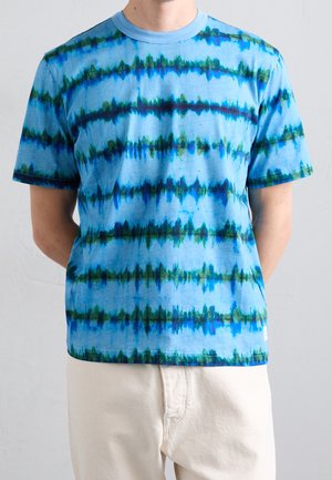 T-shirt a maniche corte con stampa tie-dye blu, con strisce orizzontali blu scuro e verdi, indossata da una persona con pantaloni beige su uno sfondo semplice.