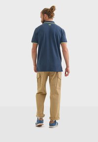 Mörkblå polo med gröna detaljer, kombinerad med khakifärgade cargobyxor och blåa träningsskor. Tyget verkar slätt, med en standard krage.