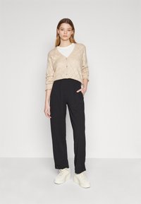 Beige knitted cardigan with diamond pattern, black high-waisted trousers, and cream platform sneakers. Model stands with hands in pockets.
