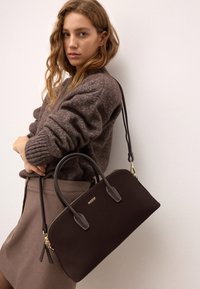 Barna nubuk táska fekete bőr részletekkel. Aranyszínű fémkeretekkel, cipzáros záródással és két felső fogantyúval rendelkezik. Világos háttér előtt elhelyezve.