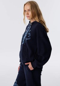 Marineblauwe hoodie met een voorzak, voorzien van een rafelige blauwe borduurontwerp en een relaxte pasvorm. Gemaakt van zachte, textuurrijke stof.