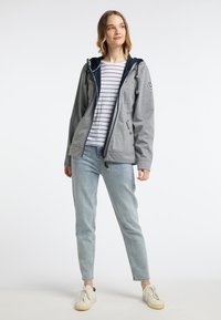 Veste zippée grise avec des accents bleu marine, capuche, cordons de serrage et poches latérales. Associée à une chemise rayée et un jean délavé. Baskets blanches.