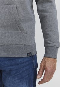 Sweatshirt gri cu dungi, având mansete ribbate și buzunar, asortat cu blugi albaștri. Eticheta vizibilă cu detalii de branding.