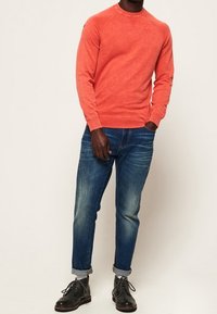 Felpa rossa con colletto rotondo e maniche lunghe, abbinata a jeans blu con risvolti e scarpe nere stringate. Presenta un logo discreto sul petto.
