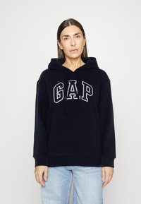 Woman wearing a navy blue GAP hoodie and light blue jeans, standing against a plain white background, looking forward with a neutral expression.