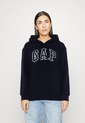 Kvinde iført en marineblå GAP hoodie og lyseblå jeans, stående foran en ensfarvet hvid baggrund, ser frem med en neutral udstråling.