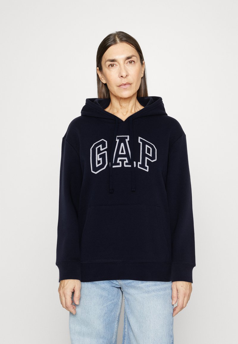 Woman wearing a navy blue GAP hoodie and light blue jeans, standing against a plain white background, looking forward with a neutral expression.