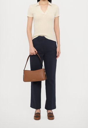 Femme portant un haut crème à manches courtes côtelé, un pantalon bleu marine, des mocassins marron, tenant un sac à bandoulière en cuir marron par sa sangle.