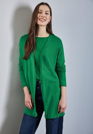 Zelený pletený cardigan s dlouhými rukávy a otevřeným předním dílem, vrstvený přes shodný zelený top. Nosený s tmavě modrými džíny, hladká textura.