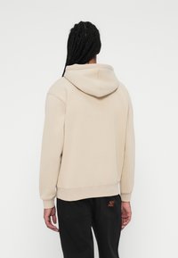 Beige hættetrøje i blødt stof med en afslappet pasform, ribbede manchetter og kant, der har et lille logo mærke på baglommen af sorte bukser.