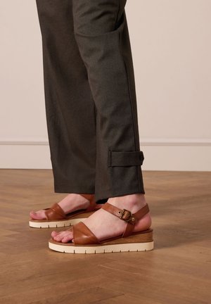 Bruine leren sandalen met brede banden en een gespsluiting om de enkel, geplaatst op een crèmekleurige, iets gestructureerde zool. Donkere broek met omslagdetails.