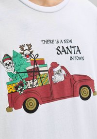 Camiseta blanca con un camión rojo, una figura de Papá Noel, un árbol de Navidad y cajas de regalos coloridas, junto a motivos de calaveras juguetonas y texto.