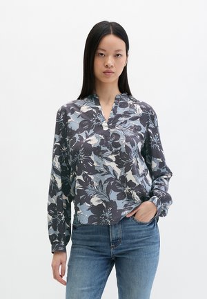 Frau trägt eine langärmelige Blumenbluse in Blau- und Grautönen, kombiniert mit blauen Jeans, stehend vor einem weißen Hintergrund.