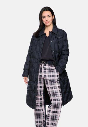 Woman wearing a long black quilted coat, black top, and pink and black plaid pants standing with one hand in her pocket.