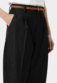 Zwarte broek met hoge taille, gemaakt van een gestructureerde stof, met zijzakken en een gevlochten bruine leren riem met een zilverkleurige gesp.