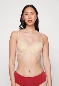 Beige pehmustettu rintaliivit säädettävillä ohuilla hihnoilla, joissa on sileä pinta ja hillitty keskimmäinen rusetti. Yhdistetty punaisiin alushousuihin.