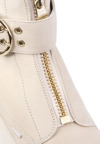 Bottine en cuir crème avec zip doré et détail de boucle, surface texturée et doublure en tissu blanc le long du zip.