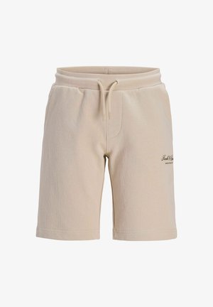 Teenager iført lyseblå t-shirt, beige shorts, hvide sokker og sort-hvide sneakers, står og smiler mens han kigger til siden.