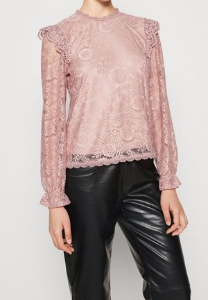 Kobieta w jasnoróżowej, kwiecistej koronkowej bluzce z falbanami na ramionach i mankietach, w połączeniu z czarnymi spodniami z ekologicznej skóry.
