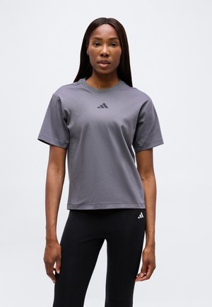 Kvinde med langt mørkt hår iført en grå Adidas T-shirt og sorte Adidas leggings, stående mod en ensfarvet lys baggrund.