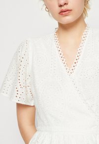 Robe blanche avec un design portefeuille, des manches courtes bouffantes, des motifs œillets complexes, un col festonné et un tissu texturé.