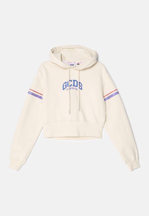 Haut court crème en tissu doux, avec un logo brodé bleu sur la poitrine et des accents rayés rouges et violets sur les manches.