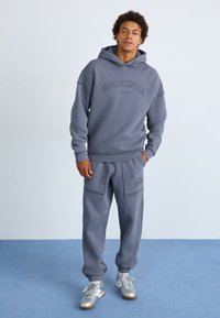Hoodie gris et joggings assortis en tissu doux, avec une coupe décontractée, des poignets côtelés et une broderie de marque à l'avant. Des baskets argentées complètent le look.