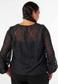 Schwarze Bluse aus einem strukturierten, transparenten Stoff mit Wellenmustern. Lange Ärmel und ein runder Halsausschnitt, der hinten mit einem Knopf geschlossen wird.