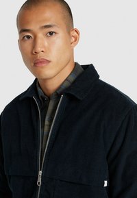Veste en velours côtelé noir avec fermeture éclair à l'avant et col, portée par-dessus une chemise à carreaux. La texture est côtelée avec un léger éclat.