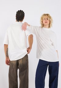 White cotton t-shirts with short sleeves, worn by two individuals, paired with dark denim pants. One shirt has a relaxed fit; the other is tucked.
