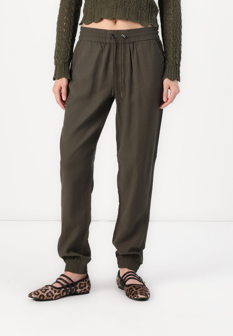 Pantalons de jogging vert olive avec des poignets et une taille élastiques, dotés d'un cordon de serrage. Associés à des chaussures plates à imprimé léopard avec des accents noirs.