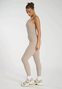 Beige bodysuit med tunna band, sömlös design och slät textur; kombinerad med vita ankelsockor och beige/svarta sportskor.