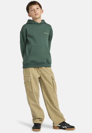 Groene sweatshirt met een voorzak en een capuchon, gecombineerd met beige cargobroek met zijzakken en zwarte sneakers met rubberen zolen.