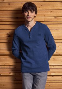 Een jonge man in een blauwe longsleeve henley shirt en grijze broek staat ontspannen tegen een wand van houten planken.