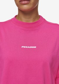 Rosa sweatshirt med hög krage och korta ärmar. Har en vit "PEGADOR" logotyp på bröstet. Mjuk, slät tygtextur.