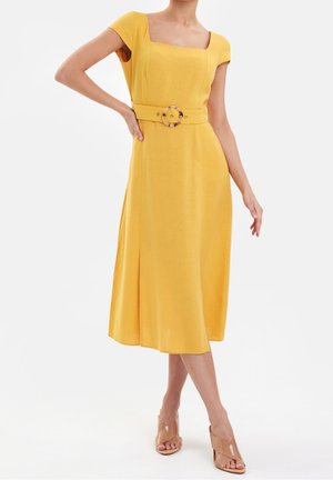 Vrouw draagt een gele midijurk met korte mouwen en een bijpassende riem met een ronde gesp, gecombineerd met beige open-teen hakken.