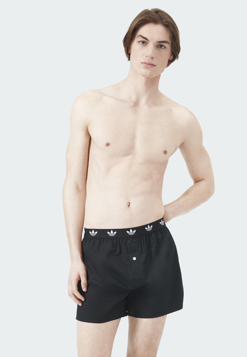 Schwarze Boxershorts aus weichem Stoff mit einem elastischen Bund, der mit weißen Trefoil-Logos gebrandet ist, und einem einzelnen Knopf vorne.