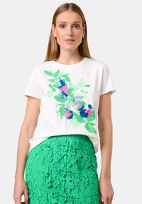 Weißes Baumwoll-T-Shirt mit grünem Blumenmuster und blauen sowie pinken Akzenten. Kombiniert mit einem grünen Spitzenrock mit floralem Muster.