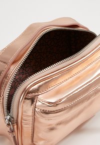 Väskan i roséguld med metallisk yta har en dragkedjestängning, slät textur och mönstrad foder med geometriska mönster.