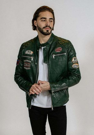Homme barbu portant une veste de course en cuir vert foncé avec des écussons, une chemise blanche et un pantalon noir, debout sur un fond uni.