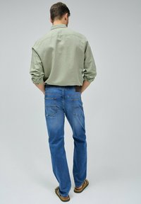 Camisa de manga larga verde claro, vaqueros azul oscuro, bolsillos visibles y una etiqueta de cuero marrón en la cintura, con sandalias marrones.