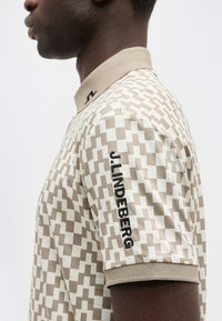 Άνδρας που φοράει μπεζ και λευκό πόλο με γεωμετρικό μοτίβο, με τη λέξη «J.LINDEBERG» κεντημένη στο μανίκι, εμφανιζόμενος σε πλάγια όψη.