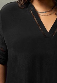 Primo piano di una persona che indossa una maglia nera a V con bordi decorativi a taglio e collane di catena argentata a strati.