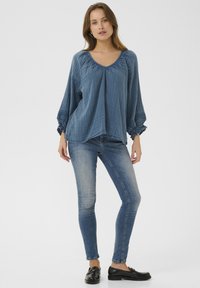 Blaues, locker sitzendes, strukturiertes Oberteil mit rundem Halsausschnitt und elastischen Bündchen, kombiniert mit engen blauen Jeans und schwarzen Slippern.