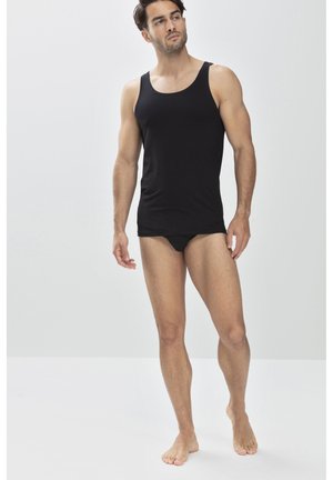 Schwarzes Tanktop aus Baumwolle. Enganliegendes Design mit breiten Trägern. Model trägt einen schwarzen Slip, steht barfuß vor einem hellen Hintergrund.