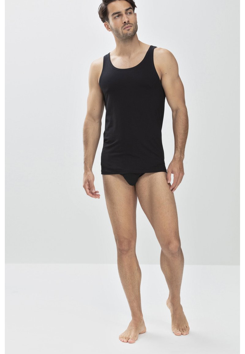 Débardeur noir en coton. Design ajusté avec de larges bretelles. Modèle portant un slip noir, debout pieds nus contre un fond clair.