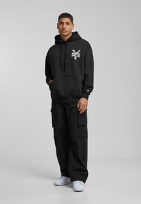 Sudadera con capucha negra y un bolsillo frontal, con un gráfico plateado. Combinada con pantalones cargo negros y zapatillas blancas.