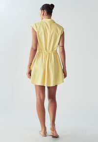 Kraega kollane kleit lühikeste varrukatega ja elastse vöökohtadega. Kangas näib pehme, voolav ja flared seelikuga. nähtavad mustrid või metallosad puuduvad.