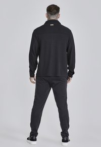 Camisa negra de manga larga con cuello y puños abotonados, combinada con pantalones ajustados a juego. El tejido parece texturizado. Calzado casual visible.